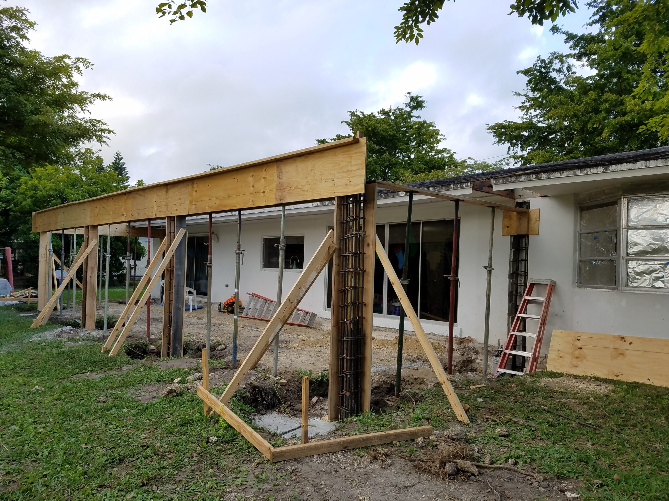 Exterior construction site of a residential patio addition featuring wooden beam formwork, exposed rebar column cages, and adjustable steel shoring jacks bracing against a single-story white house