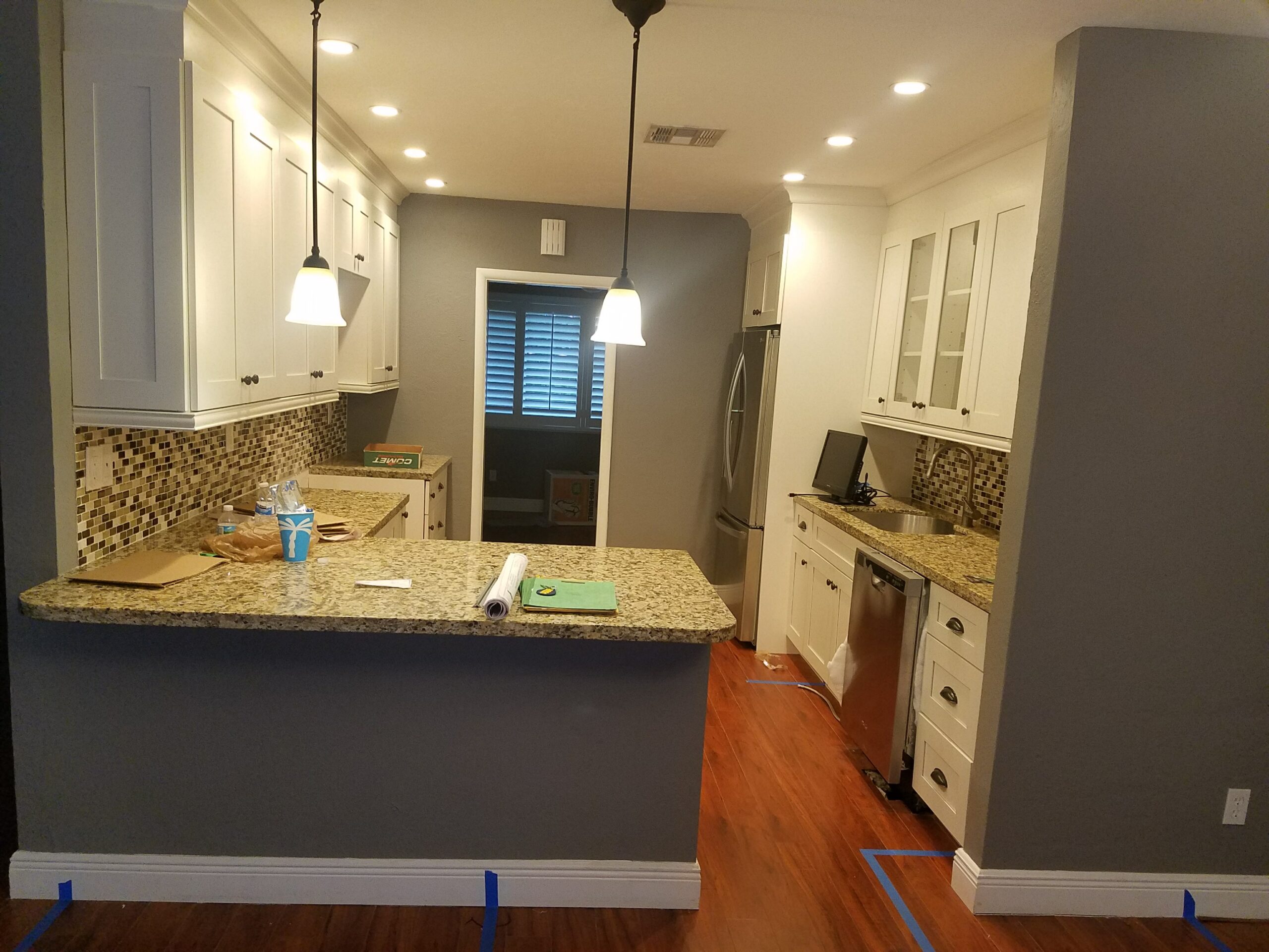 Nearly finished kitchen renovation featuring white shaker cabinets, speckled granite countertops, mosaic tile backsplash, hardwood floors, and contractor blueprints on the counter