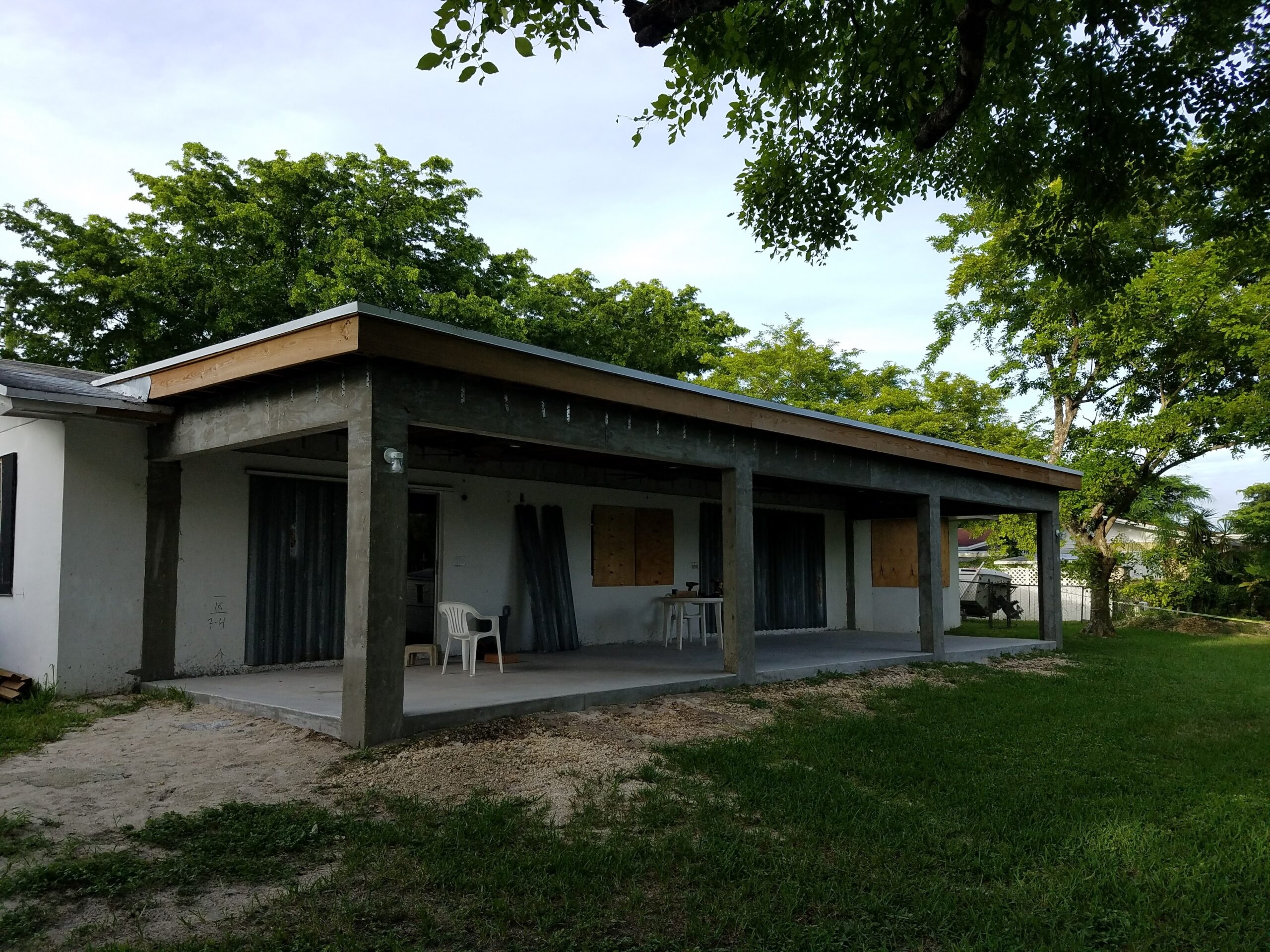 Construction progress on a residential outdoor covered patio addition featuring raw concrete support columns, a newly poured concrete slab, and wood roof framing under a clear sky