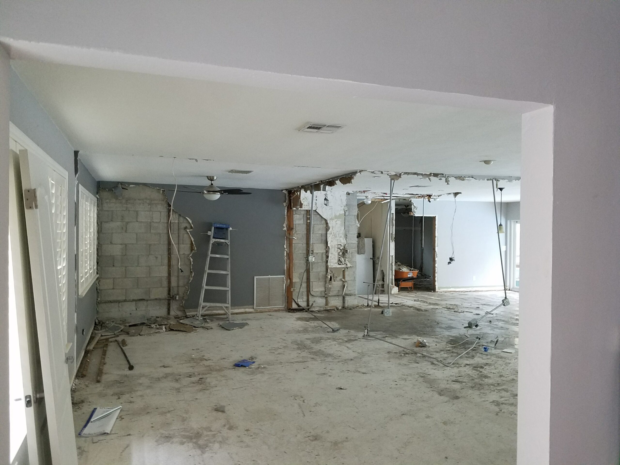 Interior home renovation site during demolition phase with partially removed drywall walls exposing concrete block, dangling electrical conduit, and bare concrete subflooring