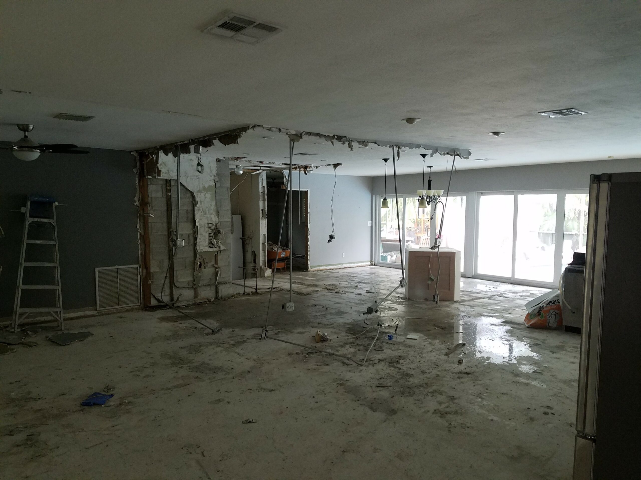 Interior home renovation during demolition stage showing exposed concrete block walls, cut ceiling with dangling electrical wires, and a bare concrete floor covered in debris and water puddles