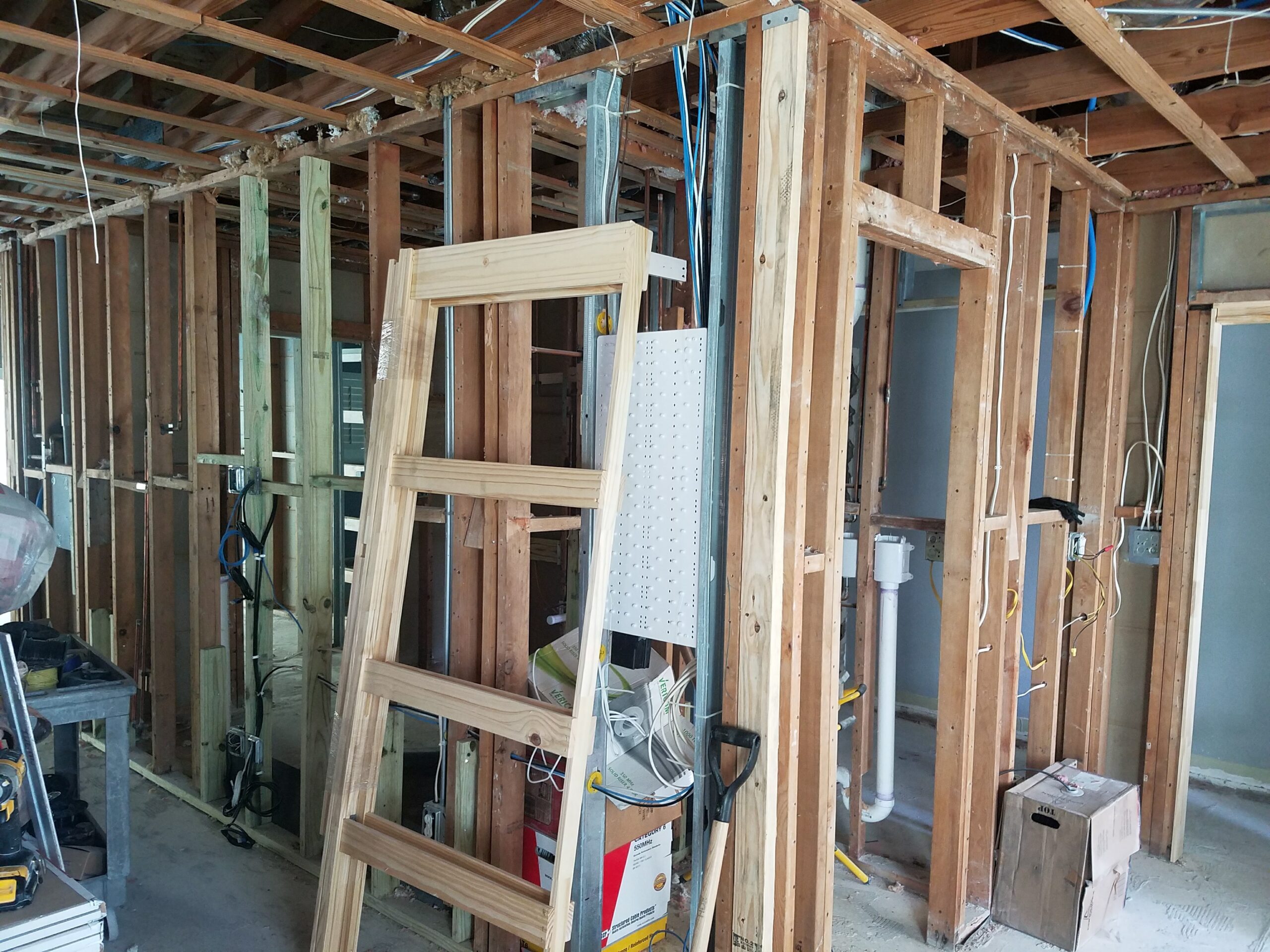 Interior construction site showing exposed wood stud wall framing, rough-in electrical and blue data wiring, PVC plumbing pipes, and a leaning wooden door frame on a concrete floor before drywall is installed