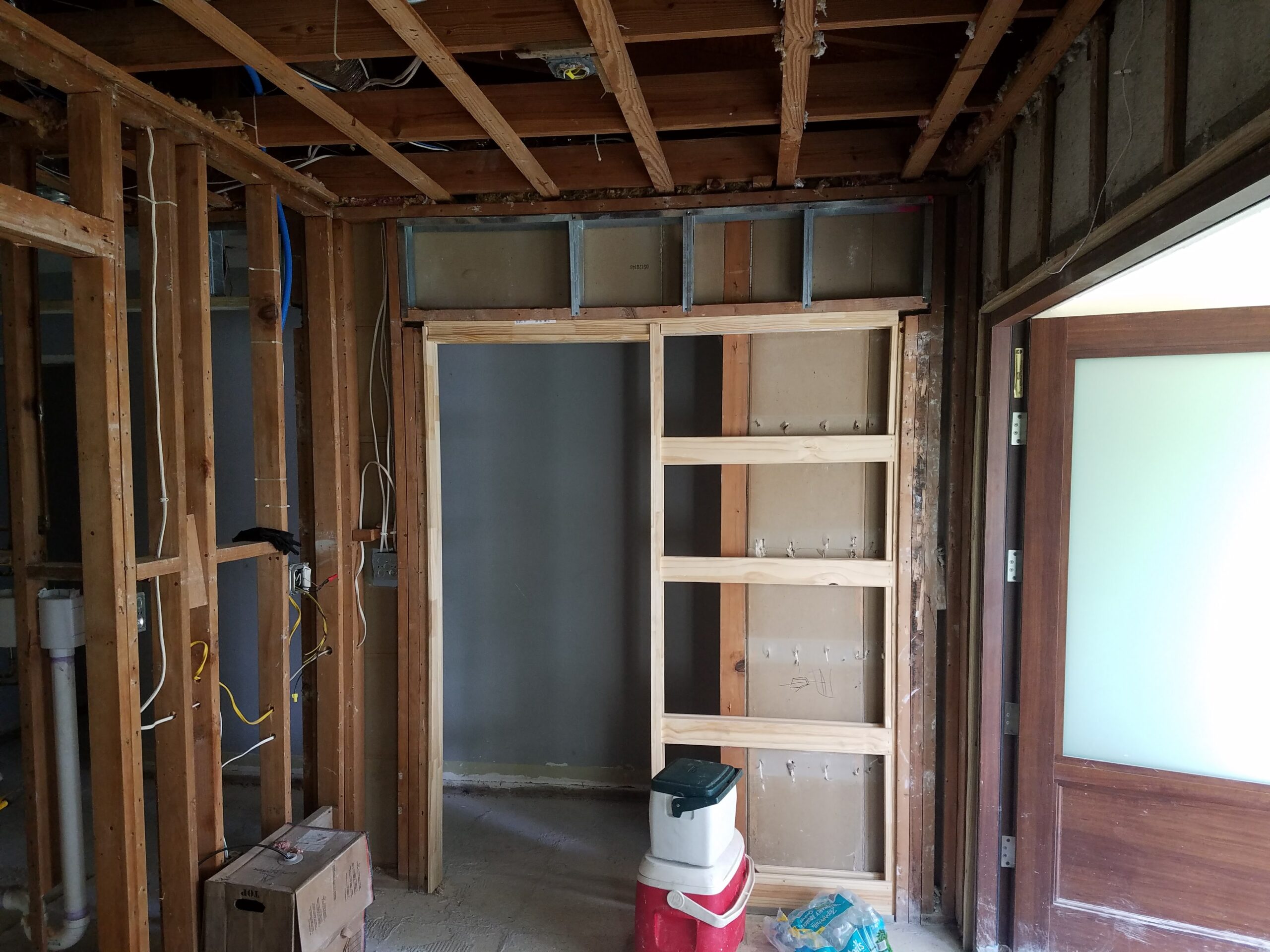 Interior residential renovation in the rough framing stage, featuring an installed wooden pocket door frame, exposed wall studs, ceiling joists, and rough-in electrical wiring