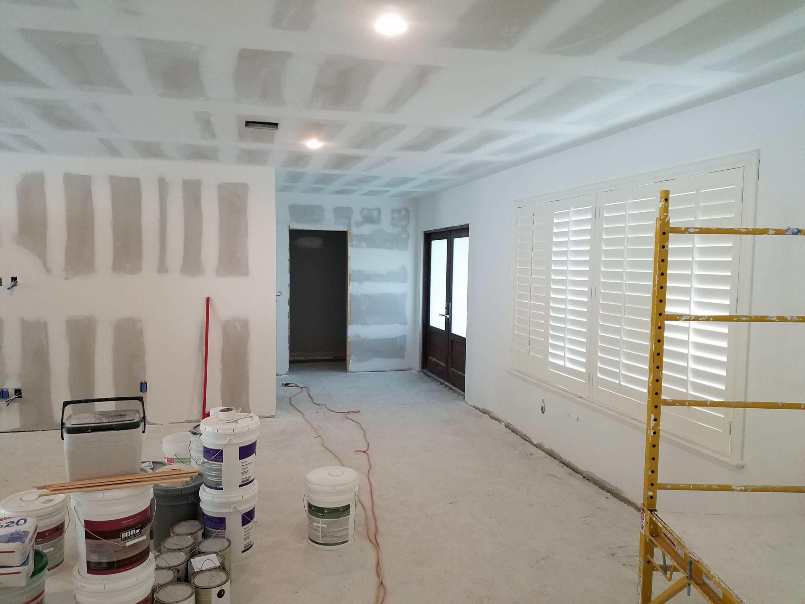 Interior home renovation site showing taped and mudded drywall seams on the ceiling and walls, with paint buckets, joint compound, and a yellow scaffolding frame on a bare concrete floor