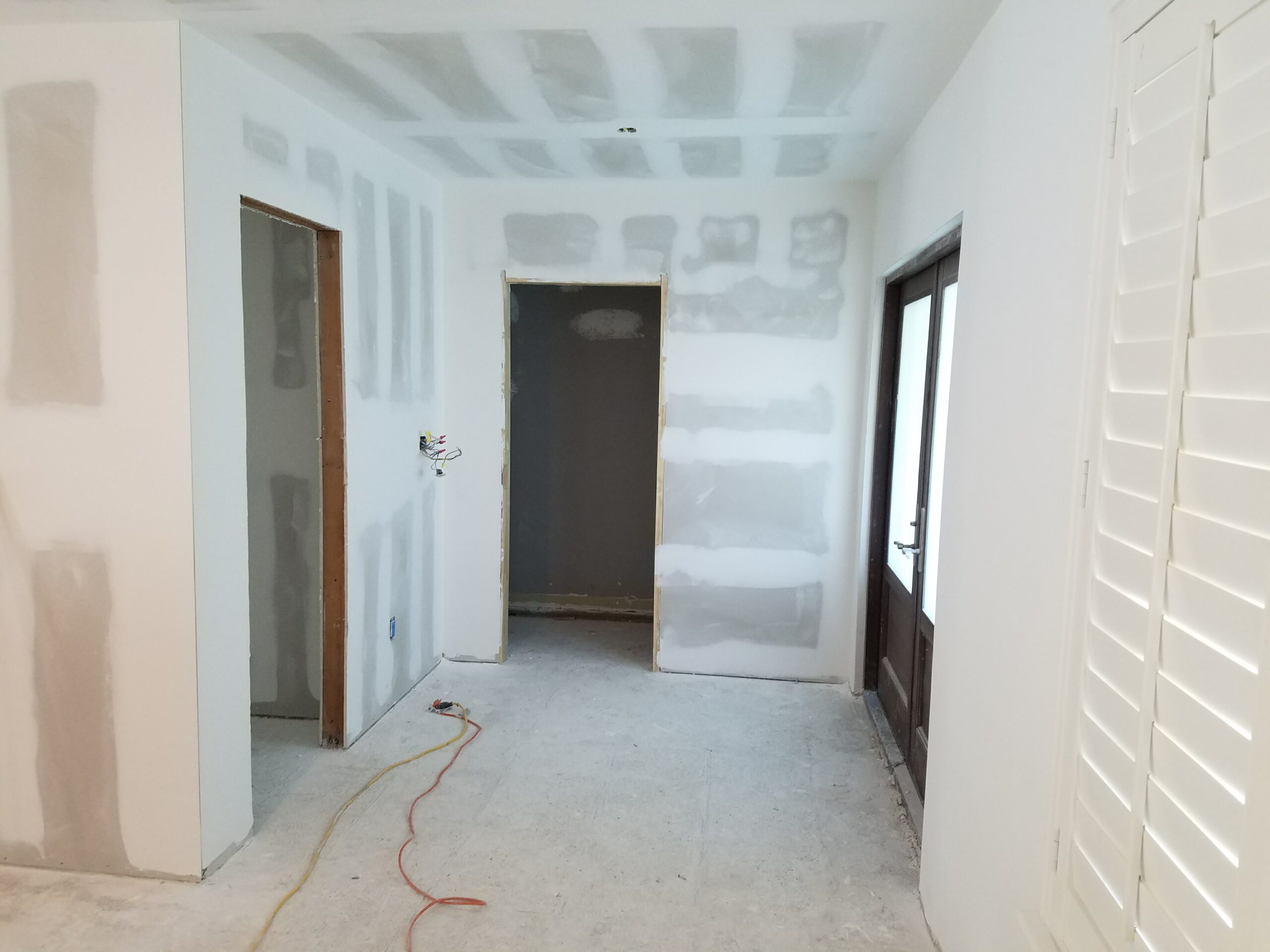 Interior room during renovation showing taped and mudded drywall on walls and ceiling, bare concrete subfloor, exposed electrical wiring, and unfinished doorways