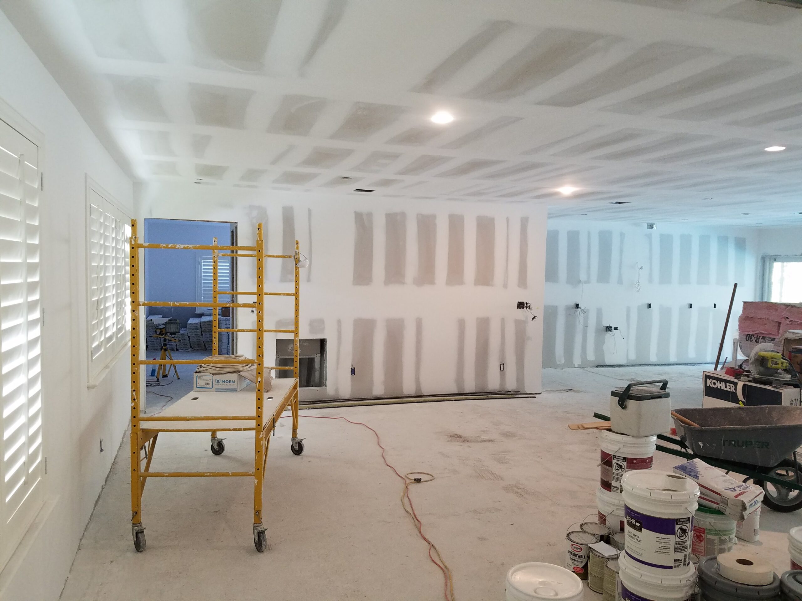 Interior residential renovation in progress showing drywall mudding and taping on the walls and ceiling of an open-concept living room, with scaffolding, paint buckets, and tools on a concrete subfloor