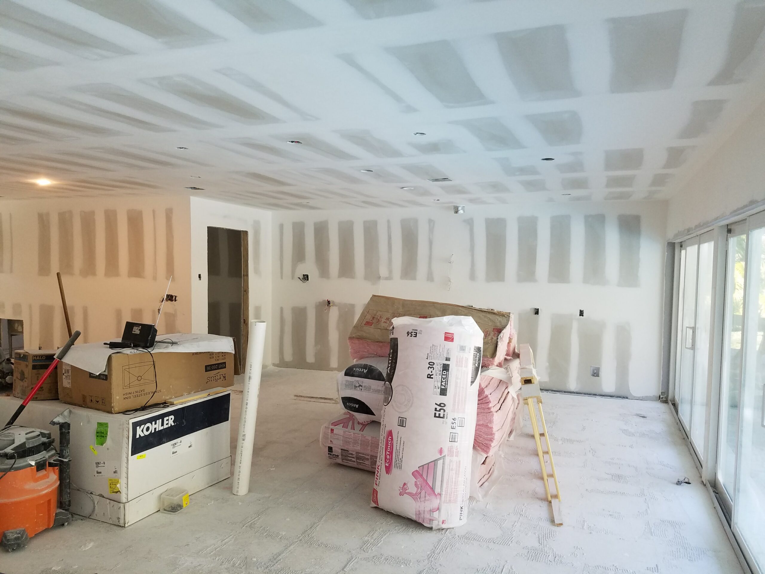 Interior residential renovation in progress showing taped and mudded drywall on ceilings and walls, stacked insulation rolls, Kohler and Kraus fixture boxes, and sliding patio doors