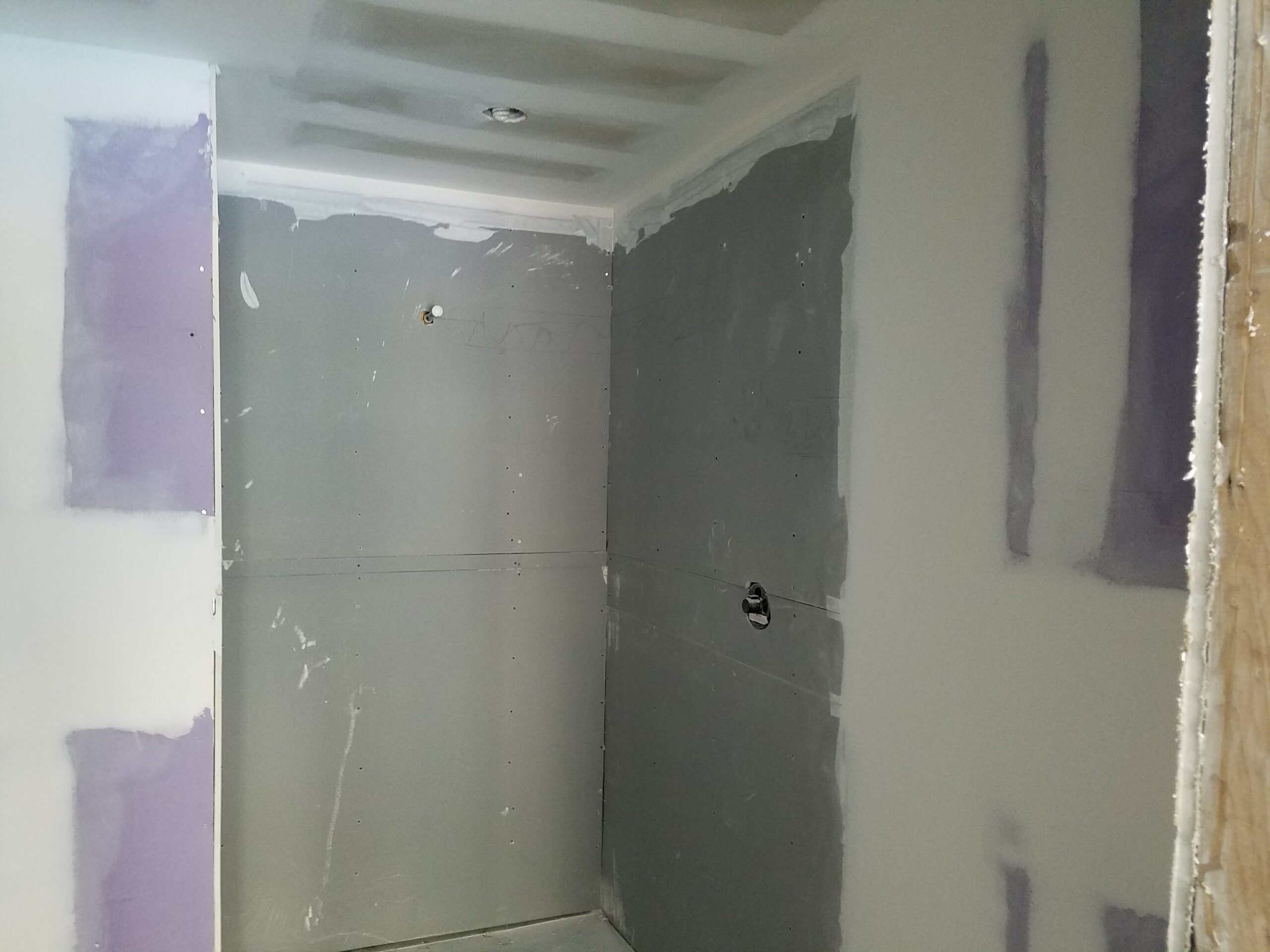 Bathroom renovation in progress showing a rough-in shower stall with installed cement backer board, taped and mudded drywall, rough-in plumbing outlets, and ceiling light fixture opening