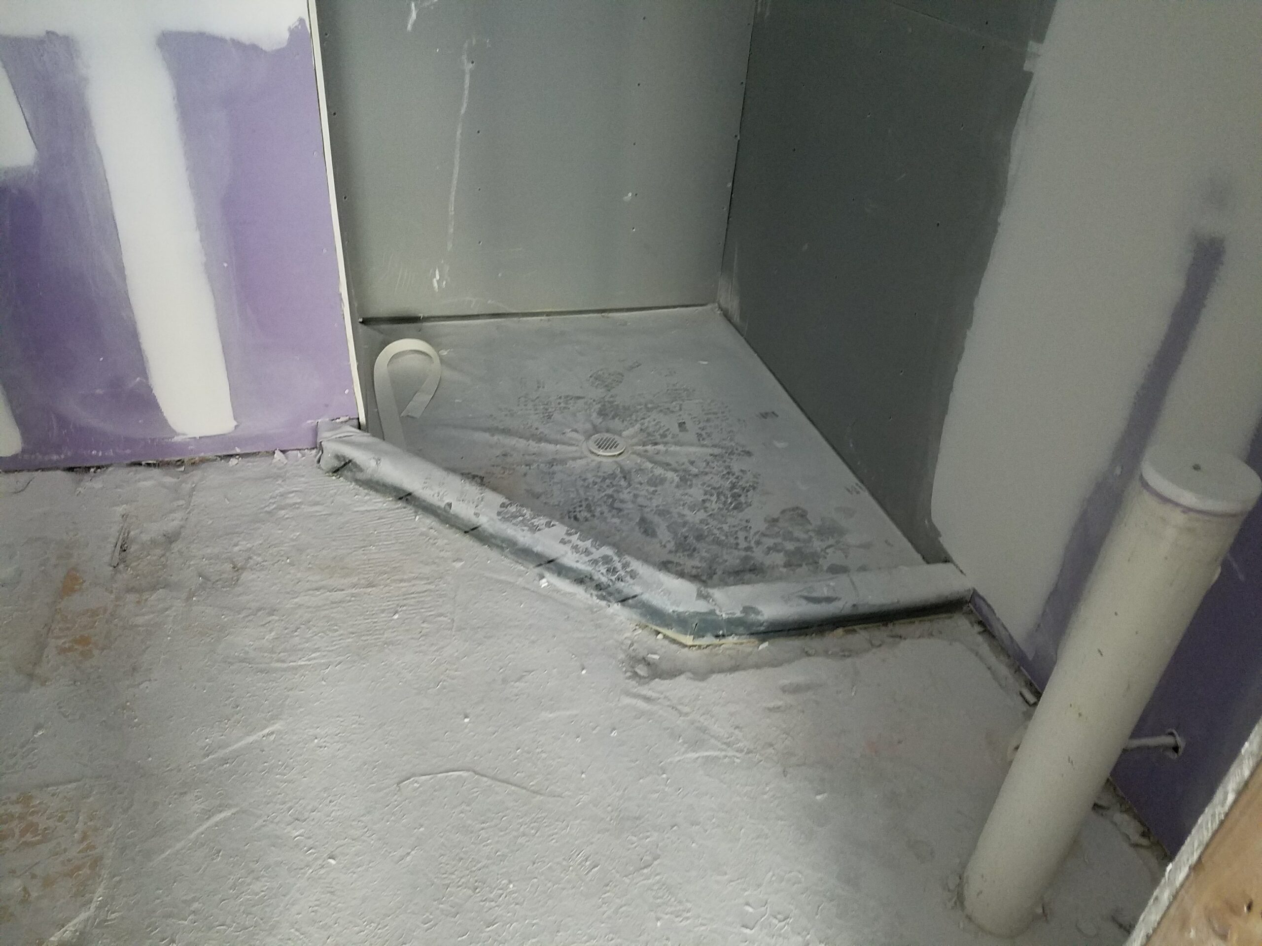 Corner shower renovation in progress showing installed sloped shower pan, drain, backer board walls, and taped purple moisture-resistant drywall before tile installation