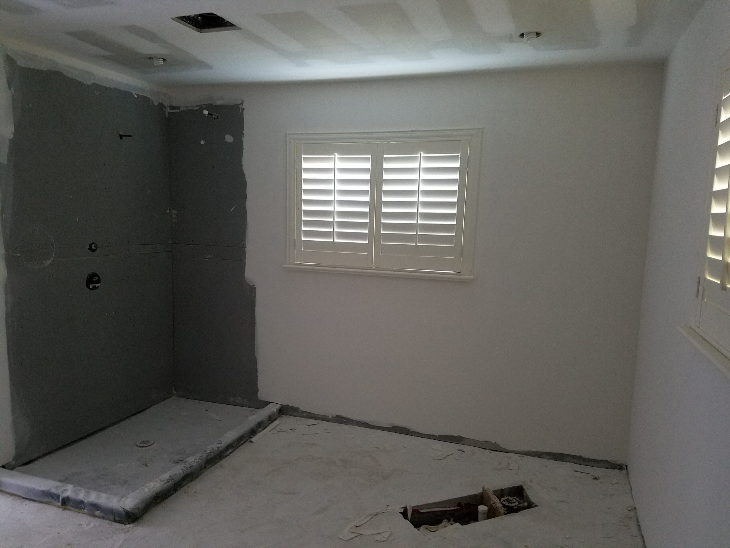 Mid-renovation view of a primary bathroom showing drywall installation, rough-in plumbing, plantation shutter window, and a waterproofed walk-in shower pan ready for tile