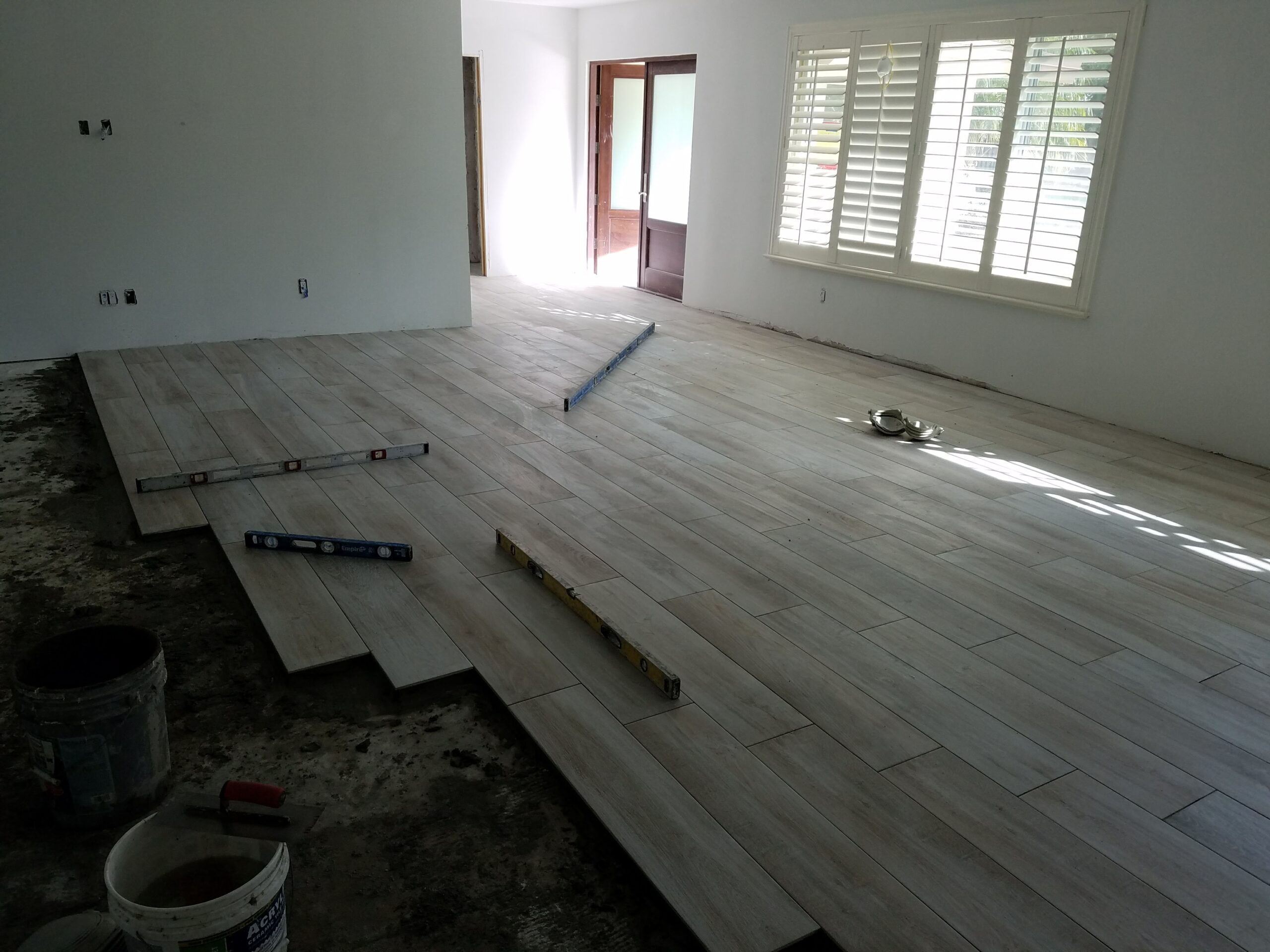 Mid-renovation view of a bright living room during the installation of light wood-look plank floor tiles, featuring levels and trowel buckets on an exposed concrete subfloor