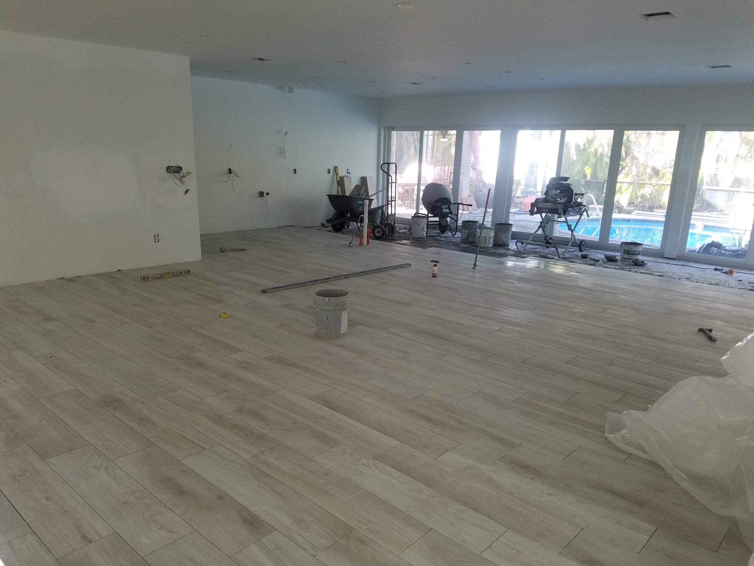 In-progress interior home renovation showing newly laid light wood-look tile flooring, tile saws, mixing equipment, and sliding glass doors opening to a backyard swimming pool