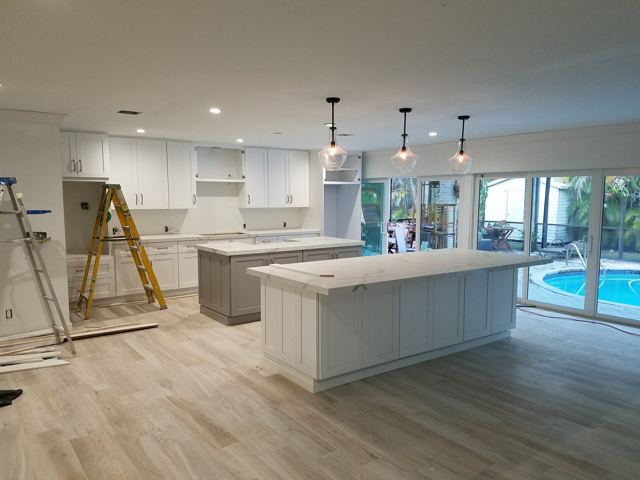 Late-stage modern kitchen remodel showing installed double islands with white quartz countertops, white cabinetry, pendant lighting, and glass patio doors overlooking a pool during construction