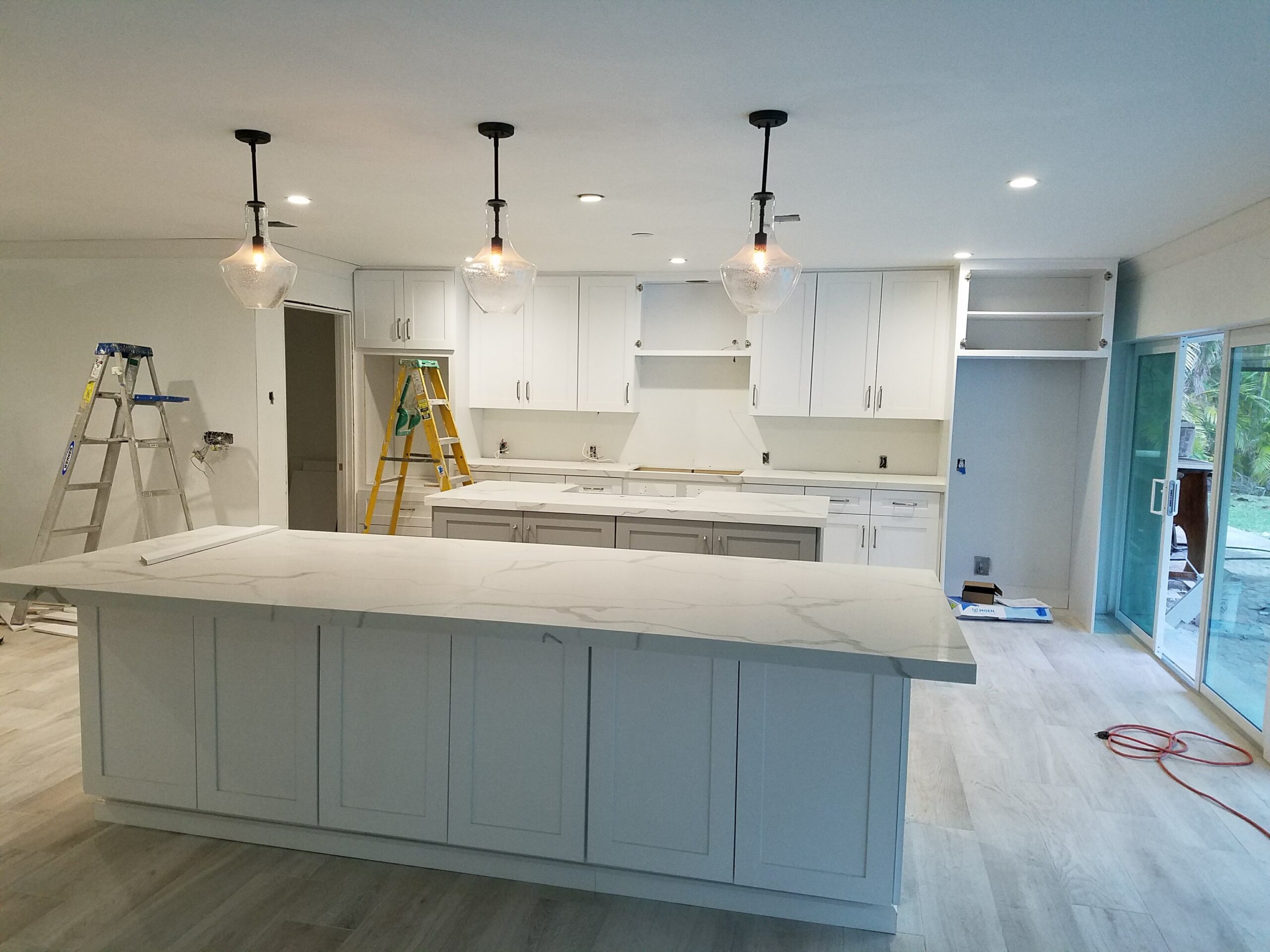 Modern open-concept kitchen under renovation with dual quartz islands, white shaker cabinets, and hanging glass pendant lights nearing completion