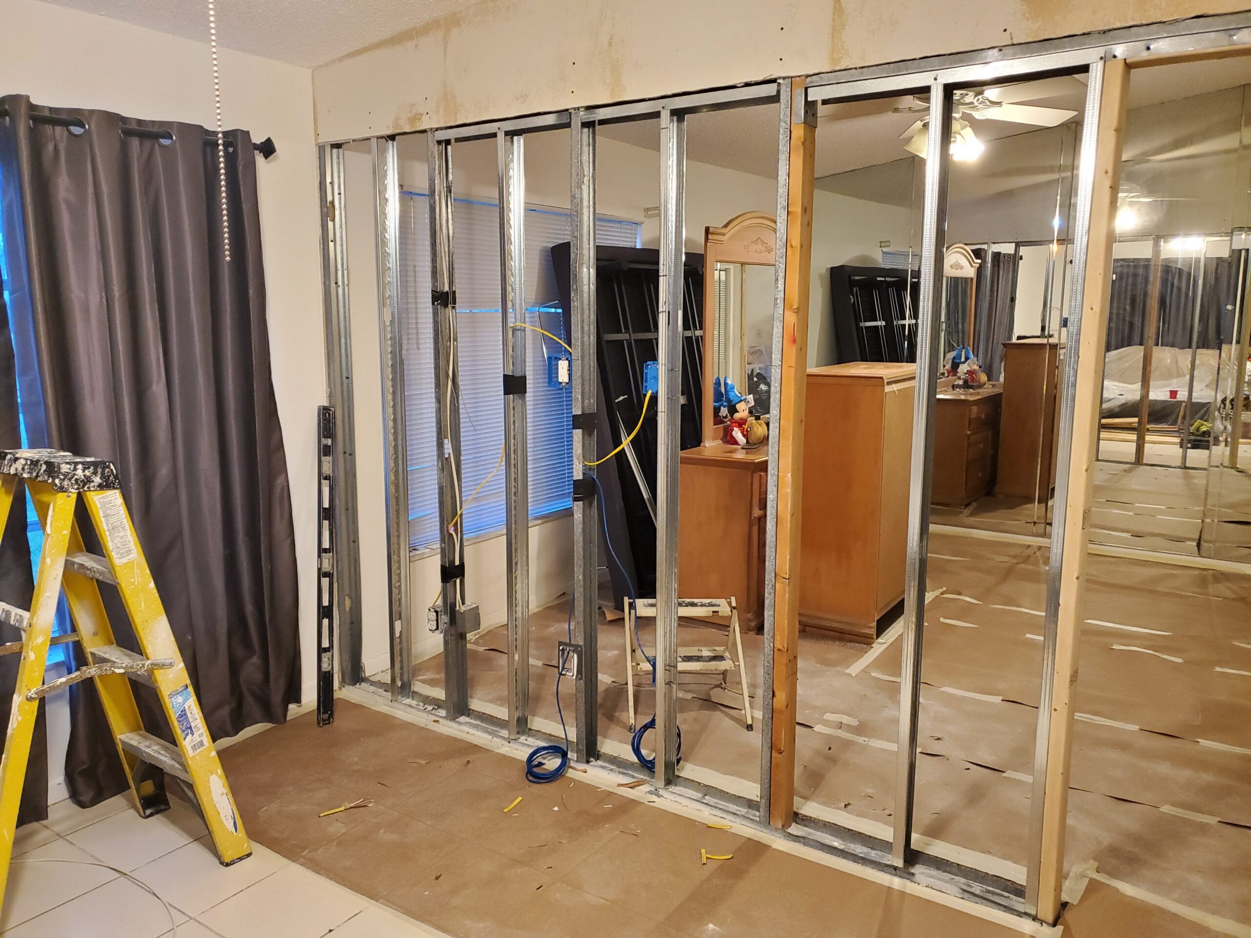 Metal stud framing for a new interior partition wall during a home renovation, featuring rough-in wiring, electrical boxes, a step ladder, and floor protection