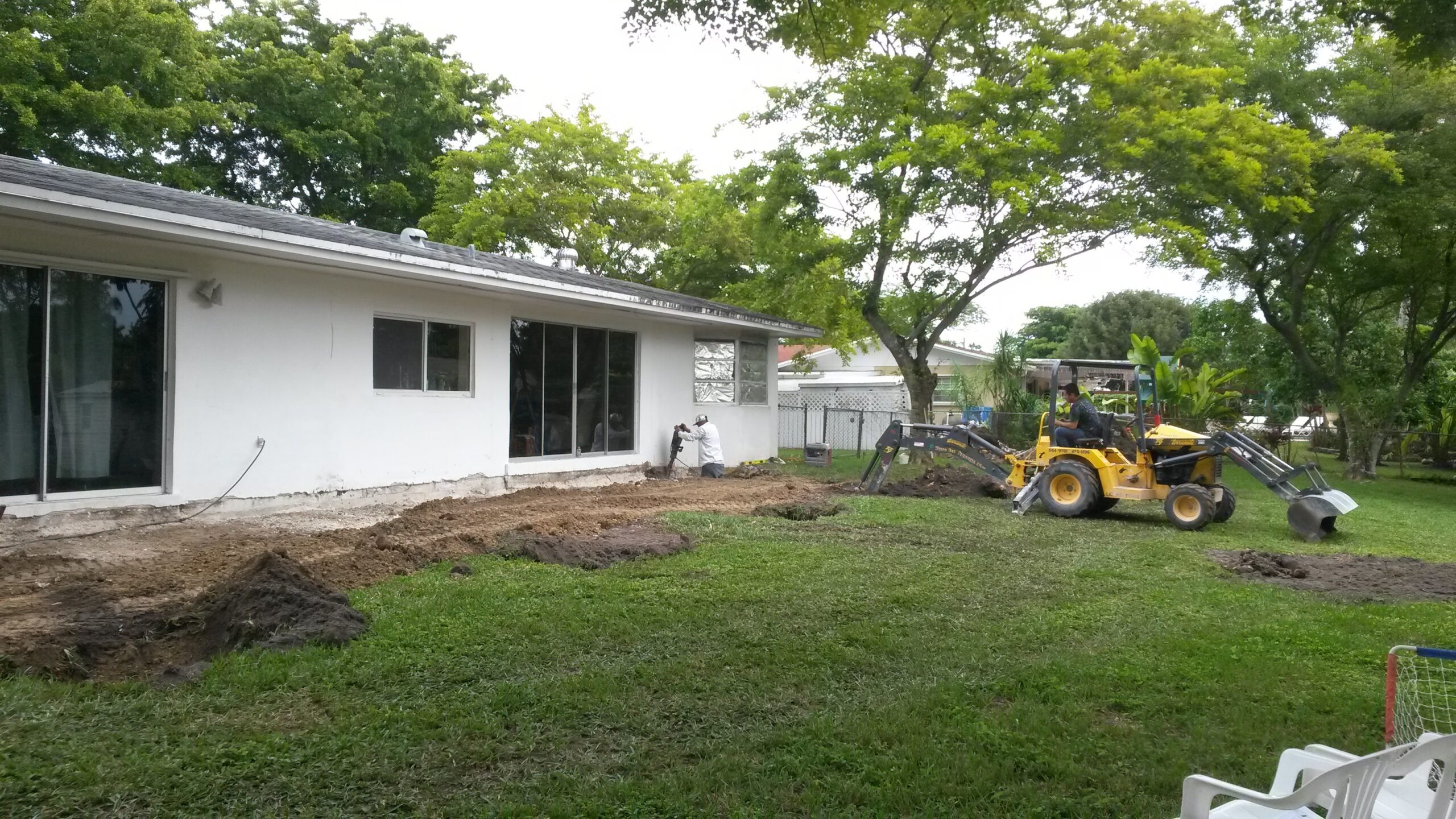Construction crew using a yellow backhoe and jackhammer to excavate soil along the rear exterior foundation of a single-story white home in preparation for an outdoor addition